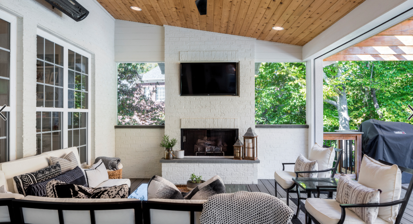 Cozy, covered patio with a fireplace, mounted television, and seating area surrounded by lush greenery, perfect for those considering home remodeling services in Charlotte.