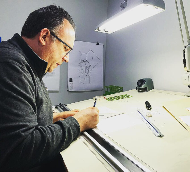 Man in glasses working on architectural blueprints at a lit drafting table, with drawing tools and a digital device nearby.