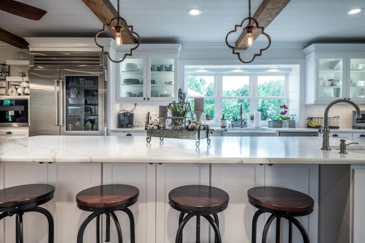 Modern kitchen with white cabinets, marble countertops, three dark wooden barstools, pendant lights, large refrigerator, and a window overlooking greenery—a perfect example of what to expect during a house remodel.