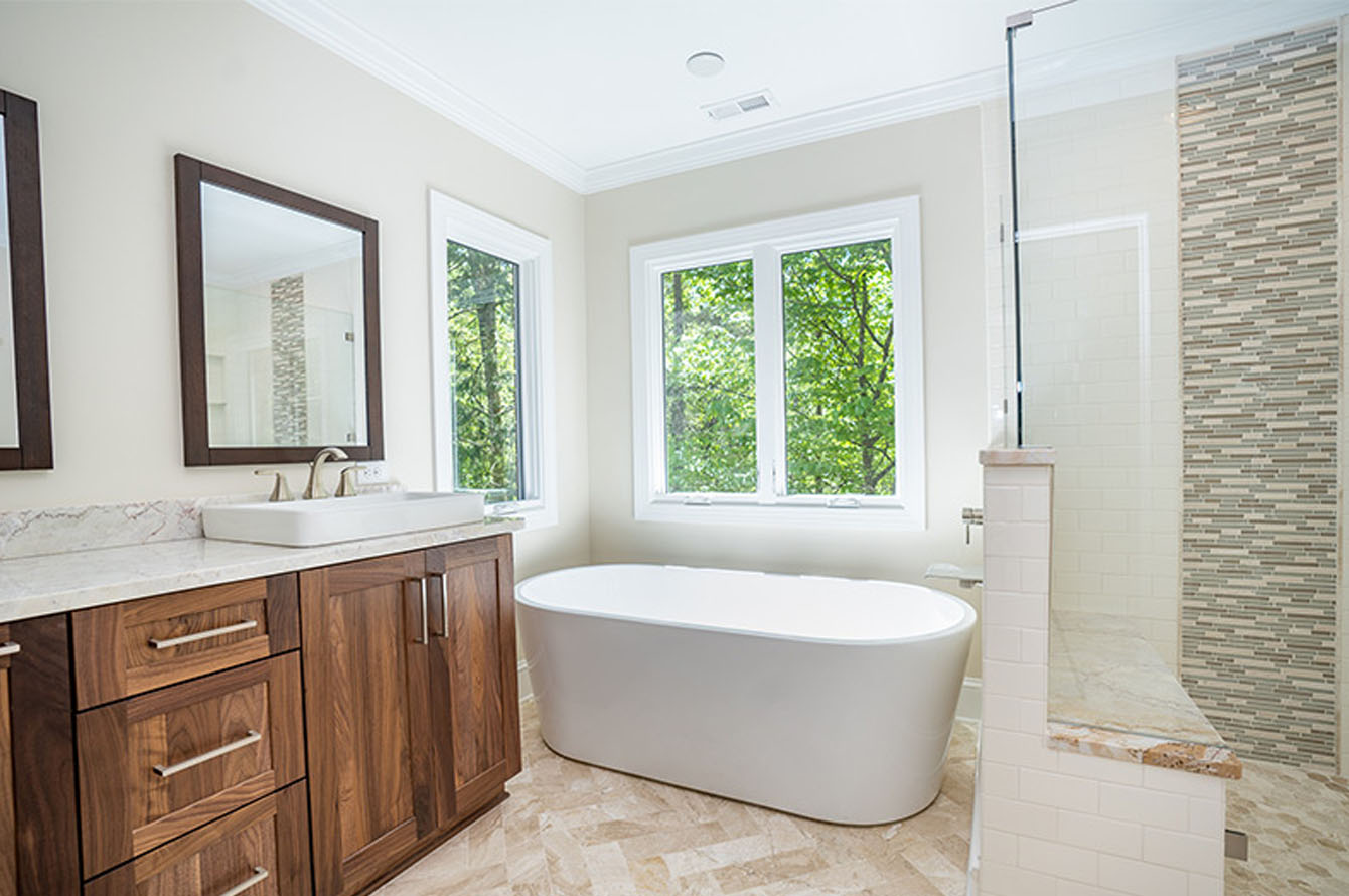 Modern bathroom remodeling with a freestanding bathtub, dual sinks, wooden cabinets, and a large window overlooking greenery.