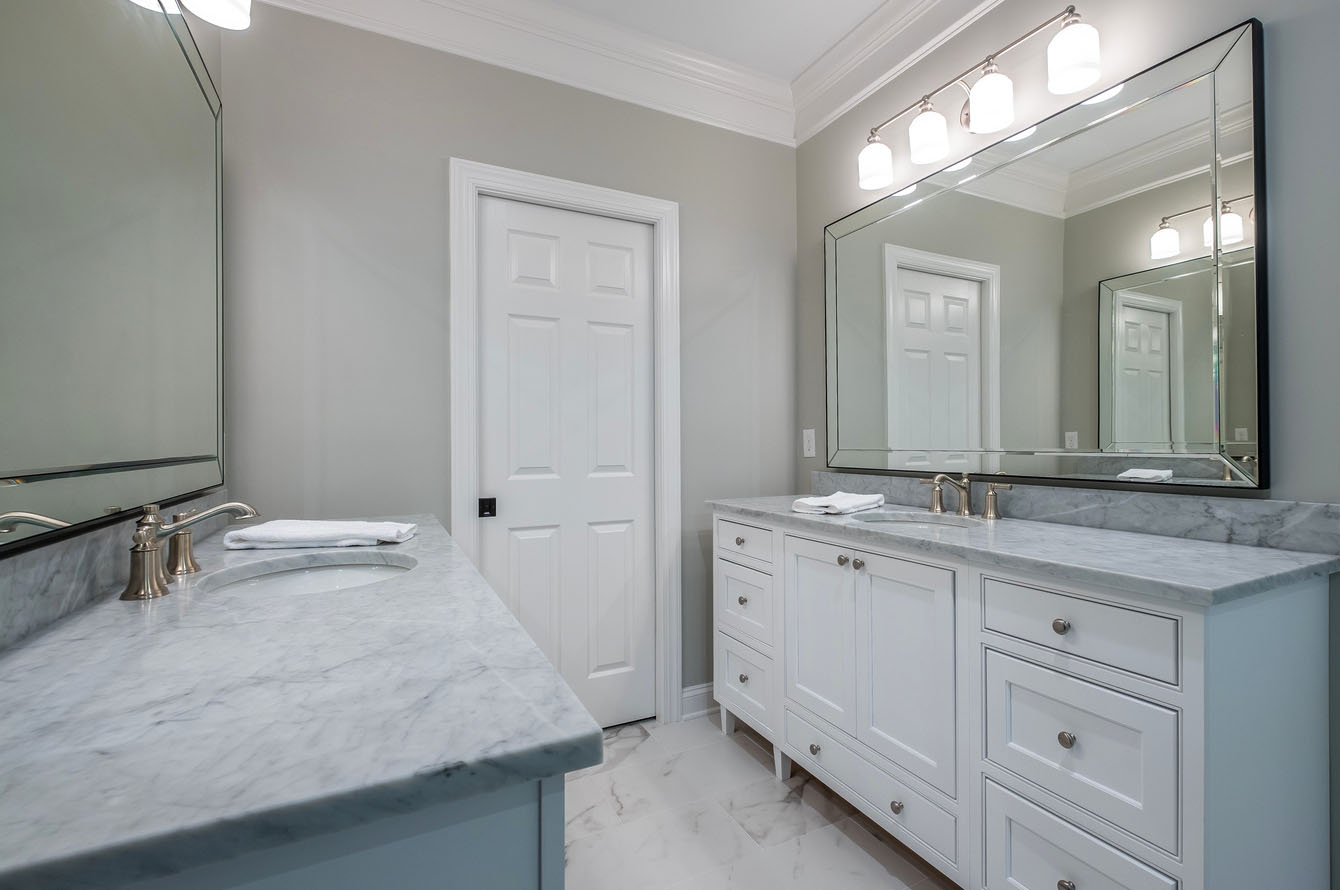 Modern bathroom remodeling featuring white cabinetry, marble countertops, and a large mirror with reflections of a white door and a second mirror.