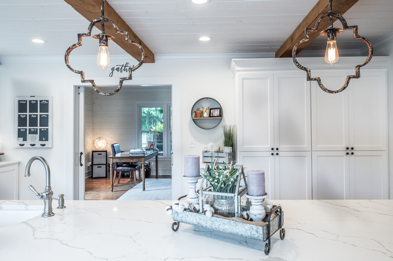 Modern kitchen remodeling with white cabinets, marble countertops, and unique pendant lights, featuring a decorative centerpiece and a view into a home office.