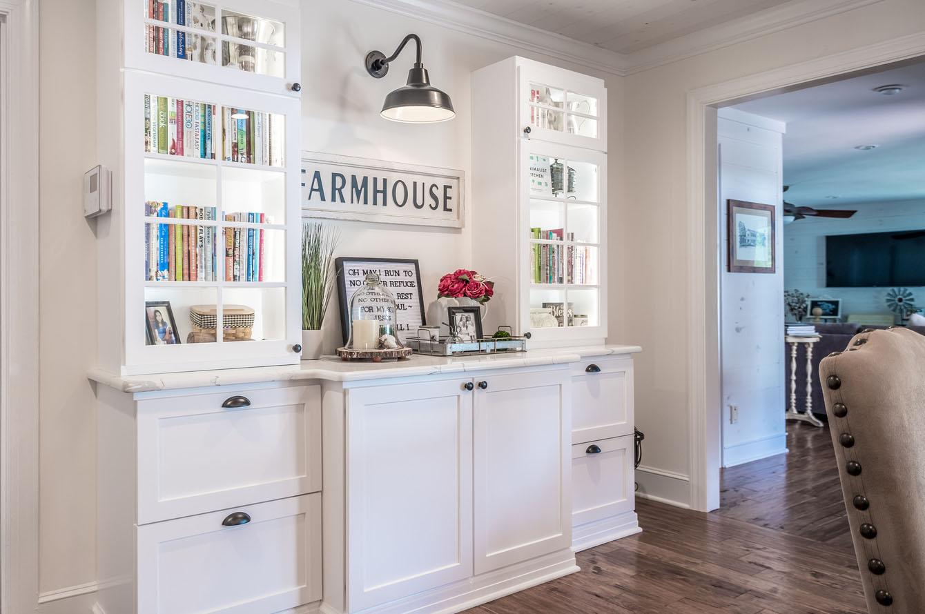 A bright, cozy corner in a farmhouse-style home featuring white built-in shelves and cabinets with books, decorative items, and flowers. There's a tufted chair and a "farmhouse" sign