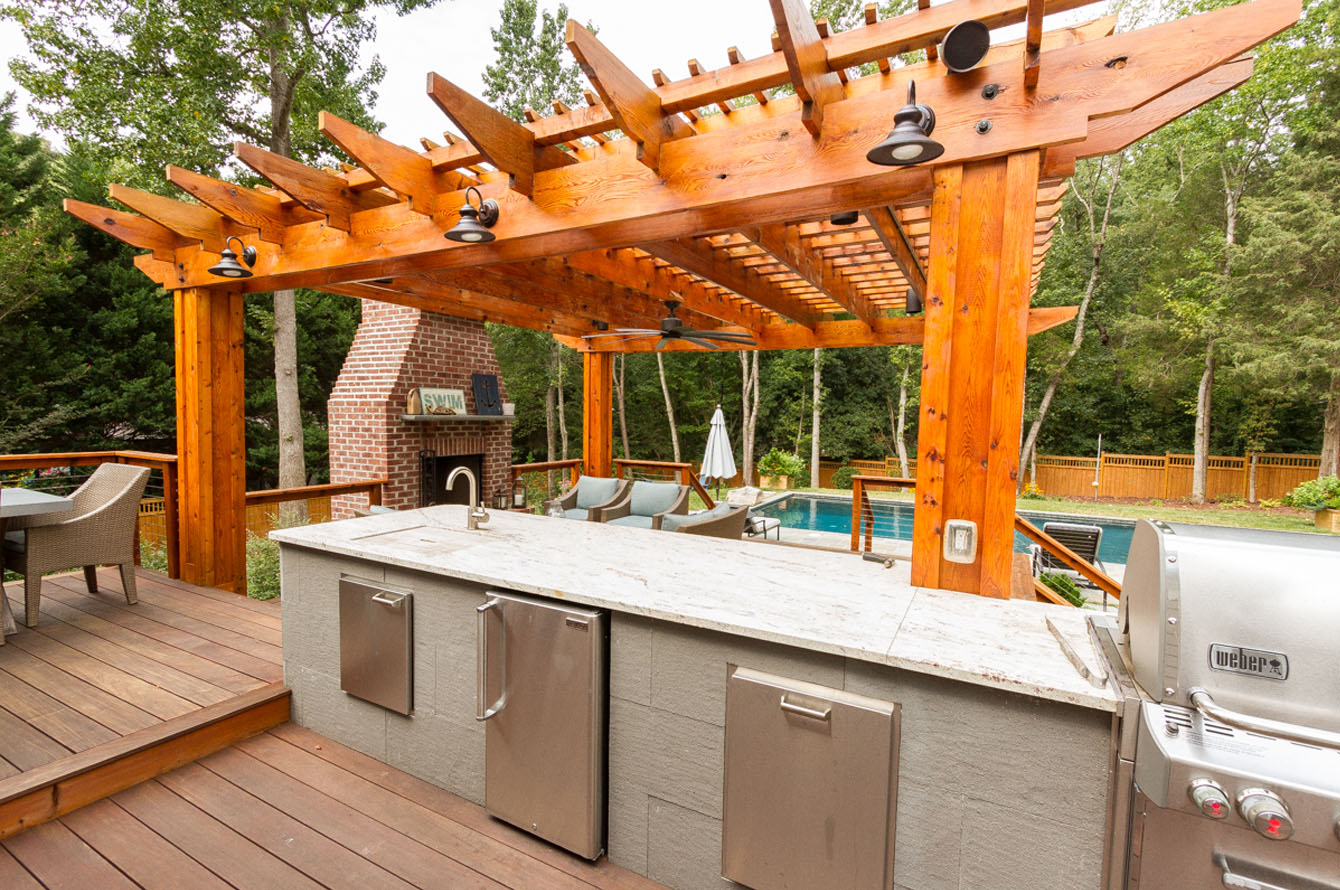 Outdoor living space featuring a kitchen with a pergola, equipped with a grill, sink, and mini-fridge, set on a wooden deck with a view of greenery.