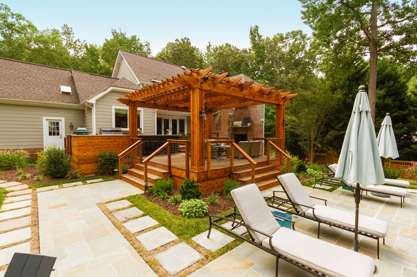 Wooden patio with pergola attached to a house, surrounded by a landscaped yard and stone pathways, featuring lounge chairs and closed umbrellas, ideal for outdoor living.