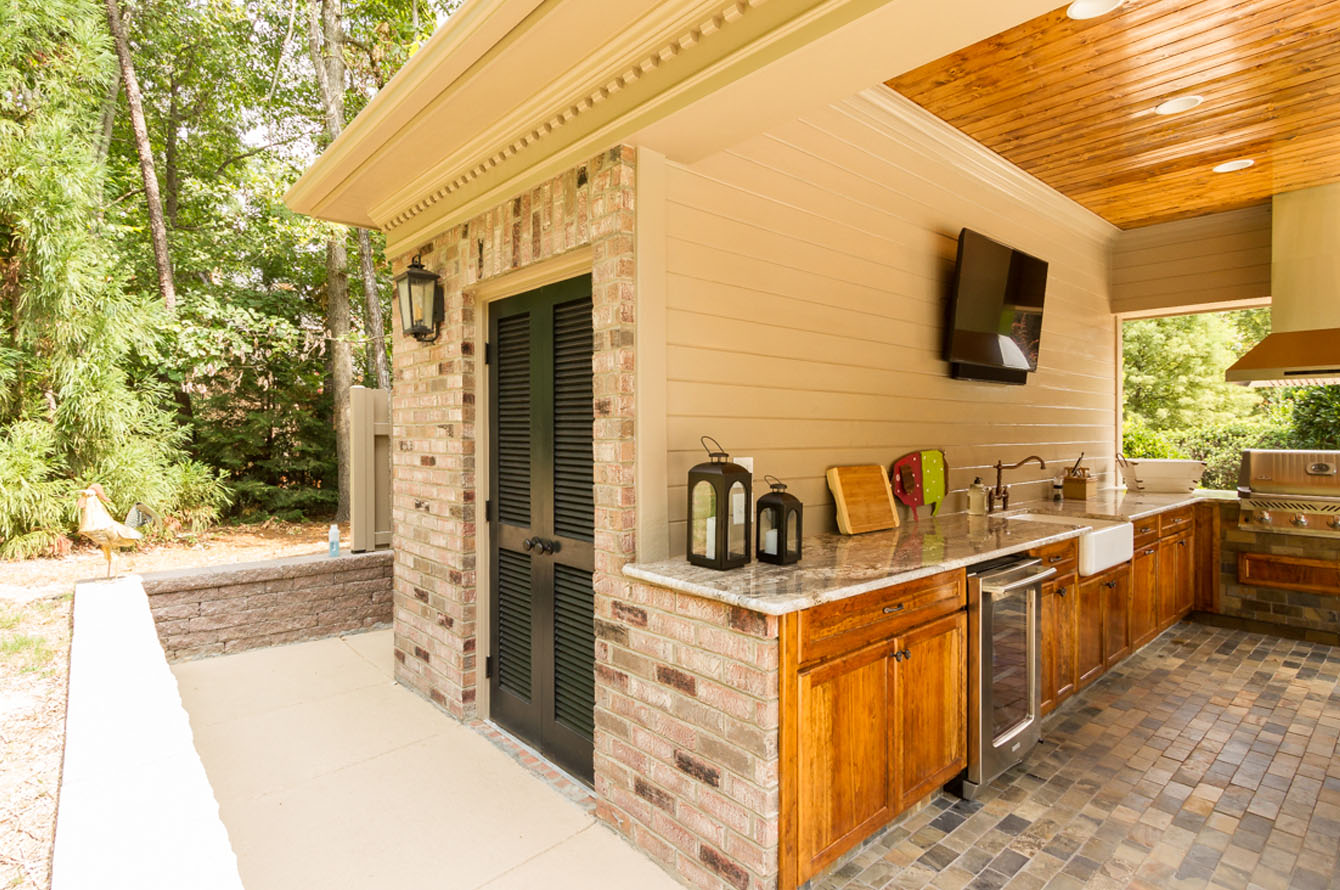 An outdoor living kitchen with wooden cabinets and a stone countertop, featuring stainless steel appliances and a TV mounted on a brick wall.