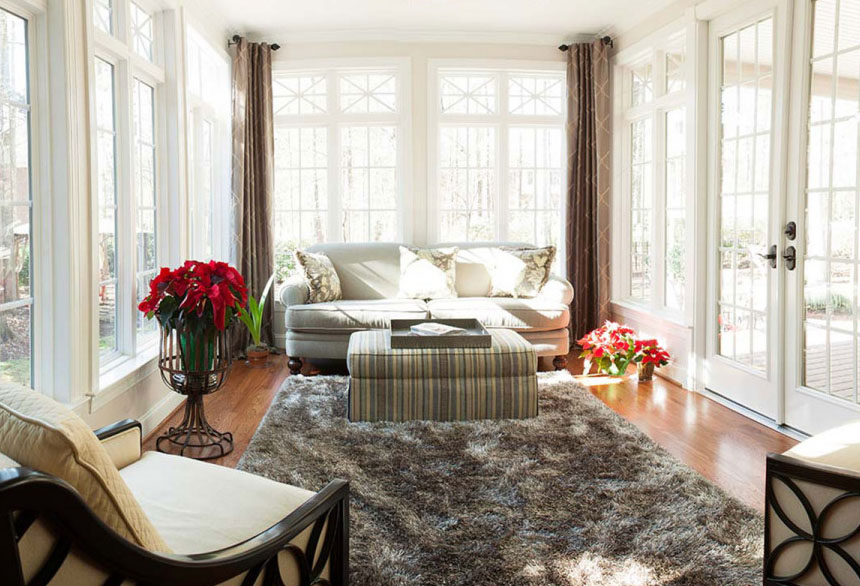 Bright, cozy sunroom with large windows, plush sofa, and a shaggy rug. Red flowers add a splash of color.