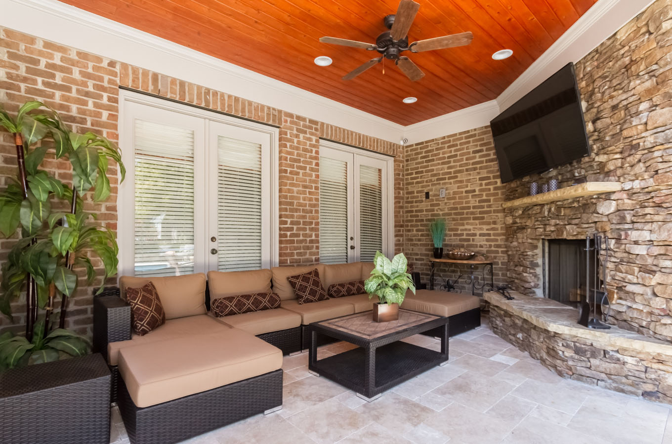 Elegant sunroom featuring a wicker seating set with cushions, a wooden ceiling with a fan, brick walls, fireplace, and a mounted television.