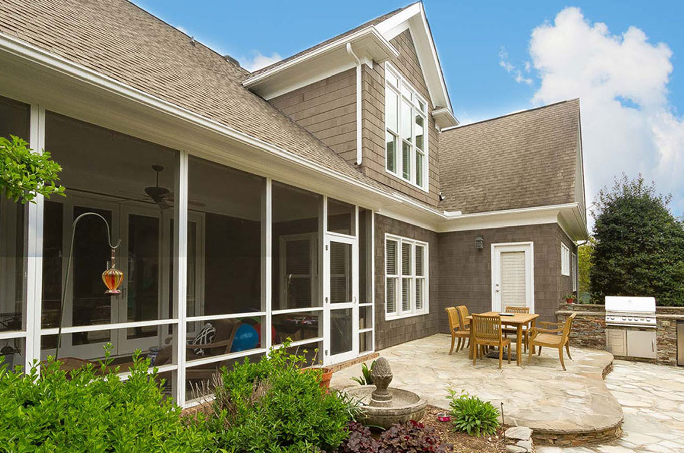 Modified Description: Modern house with a screened sunroom and a patio containing a dining table and a barbecue grill, surrounded by lush greenery under a clear sky.