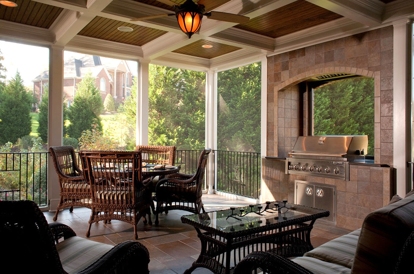 An outdoor covered patio with wicker furniture, a glass table, a built-in barbecue grill, and a fireplace, overlooking a garden has been transformed into one of the cozy sunrooms.