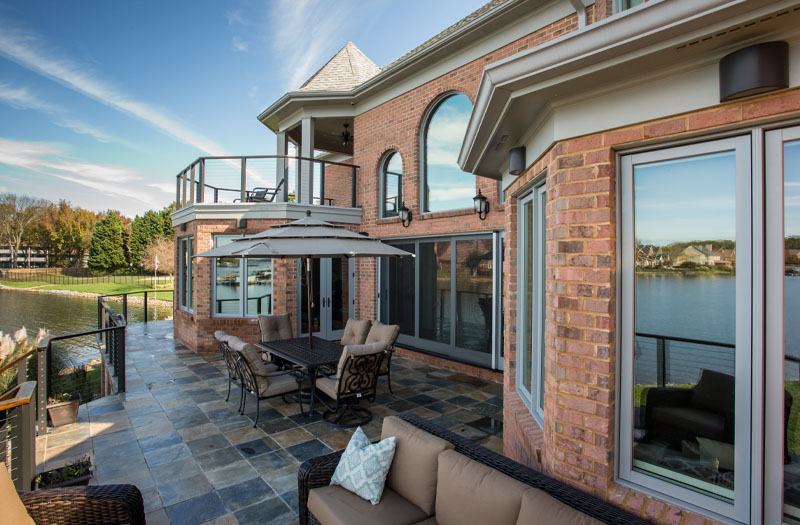 A luxurious brick house with a large patio, outdoor living furniture, and views of a lake under a clear sky.