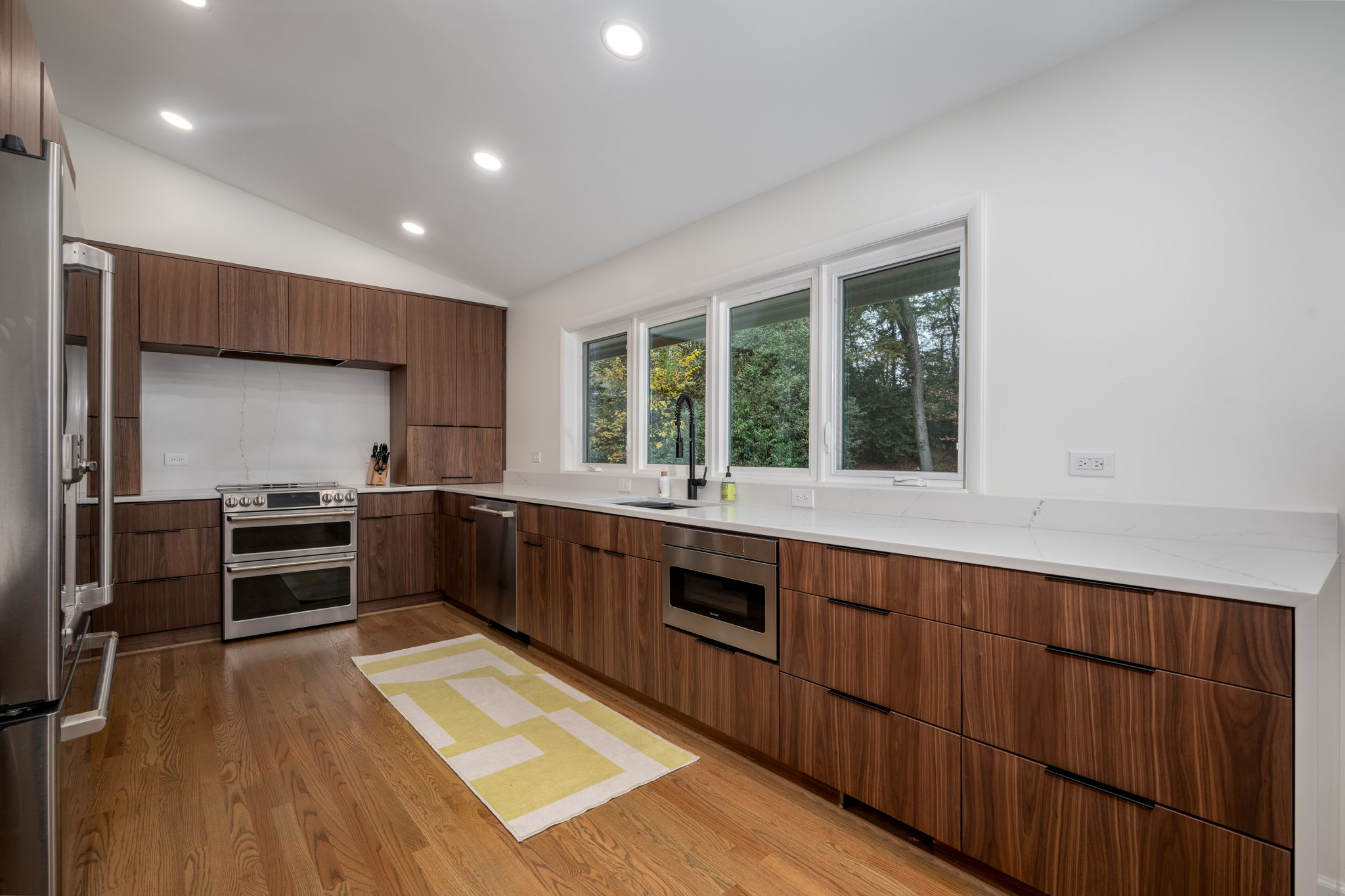 Modern kitchen, one of the most popular remodeling projects, with wooden cabinets, stainless steel appliances, and white countertops. A green and yellow rug is on the wooden floor near the sink.