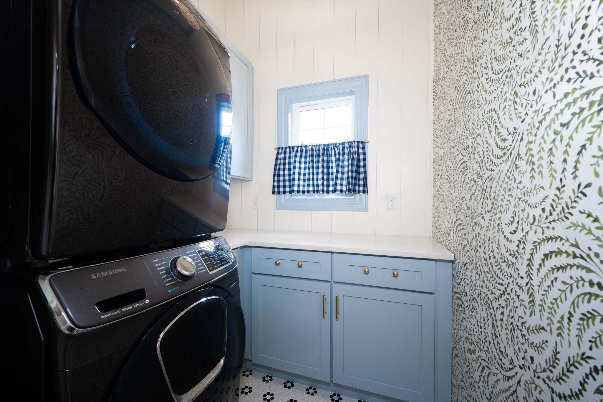 A small laundry room with a black washer and dryer, blue cabinetry, and walls covered with green leafy wall coverings.