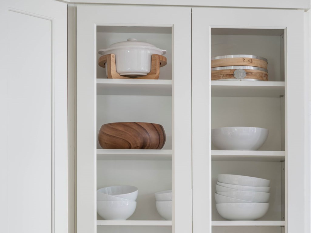 A kitchen cabinet with glass doors, displaying white and wooden bowls, a white pot with a wooden holder, and stacks of white dishes on three shelves.