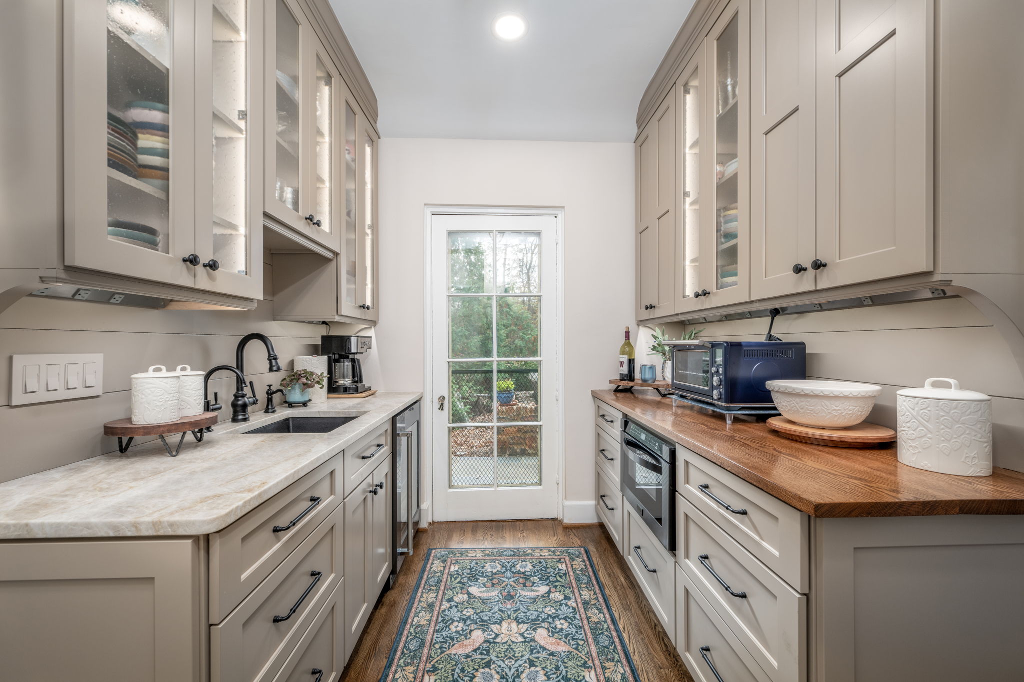 A narrow kitchen with beige cabinets, marble countertops, a small sink, stove, microwave, and various kitchen utensils. A patterned rug lies on the wooden floor, leading to a glass door at the end.