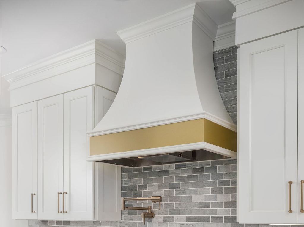 A kitchen with white cabinets, a gold-trimmed white range hood, and a gray brick backsplash. There is a stainless steel stove and a brass faucet.