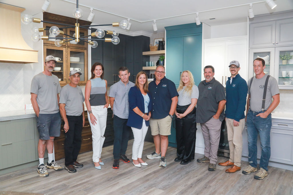 A group of nine people standing in a kitchen showroom, consisting of both men and women casually dressed, posing for a photo. The kitchen has modern cabinetry and stylish lighting fixtures.