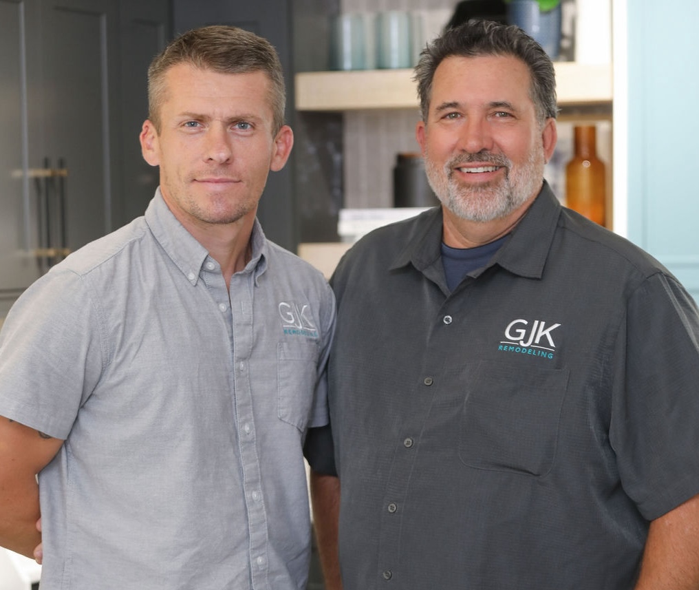 Two men wearing GJK Remodeling shirts stand shoulder to shoulder, facing the camera and smiling. They are in a modern office or kitchen setting.