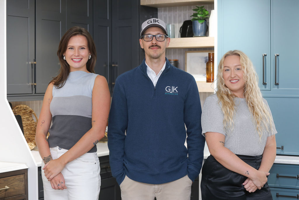 Three people are standing inside a modern kitchen showroom. The two women on either side are smiling, and the man in the center is wearing a cap and glasses. Kitchen cabinets and shelves are in the background.