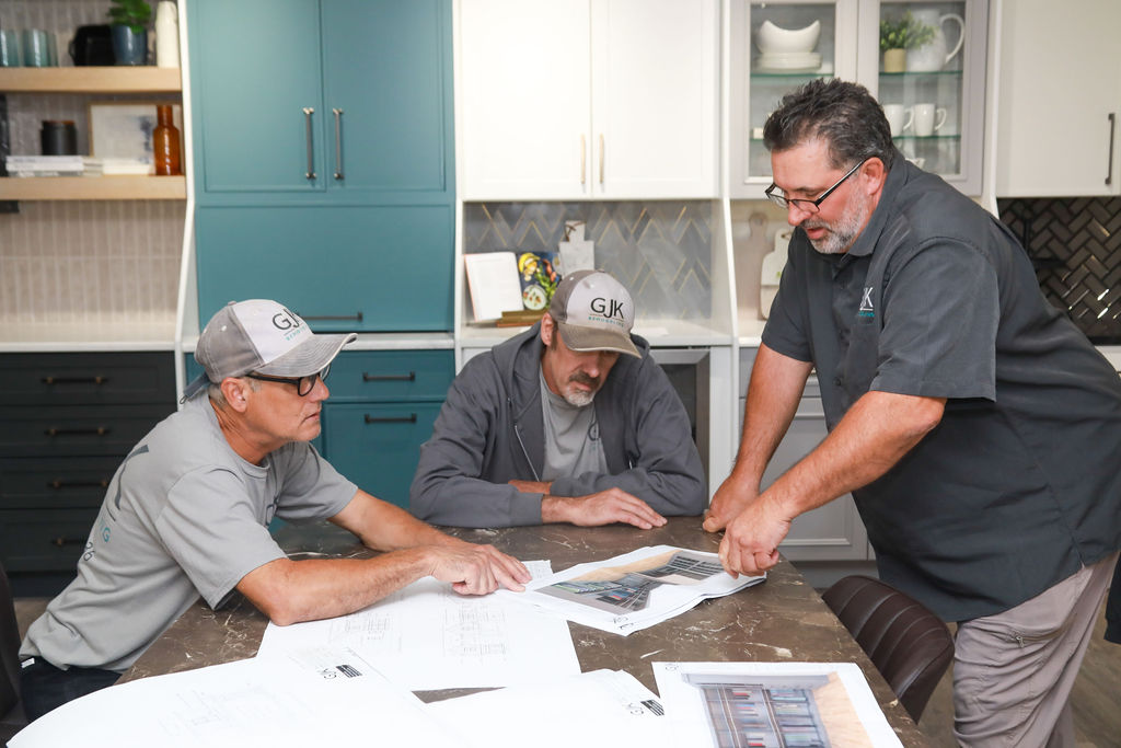 Three men in matching grey uniforms review architectural plans at a table in a modern kitchen with teal and white cabinets.