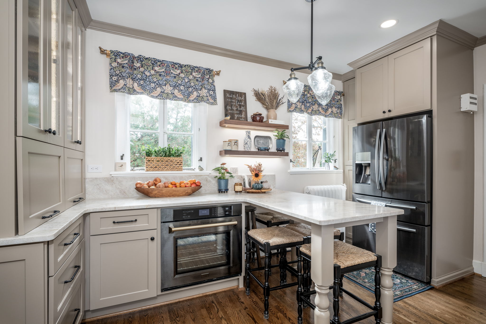 A modern kitchen with gray cabinets, stainless steel appliances, an island with seating, and large windows. The counter has a bowl of apples and various decorative items. Two pendant lights hang above the island.