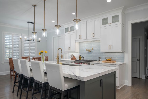 Modern kitchen with a marble island, white cabinets, and pendant lighting. Four high-back chairs, a row of hanging lights, and a dining table with chairs are visible in the background.