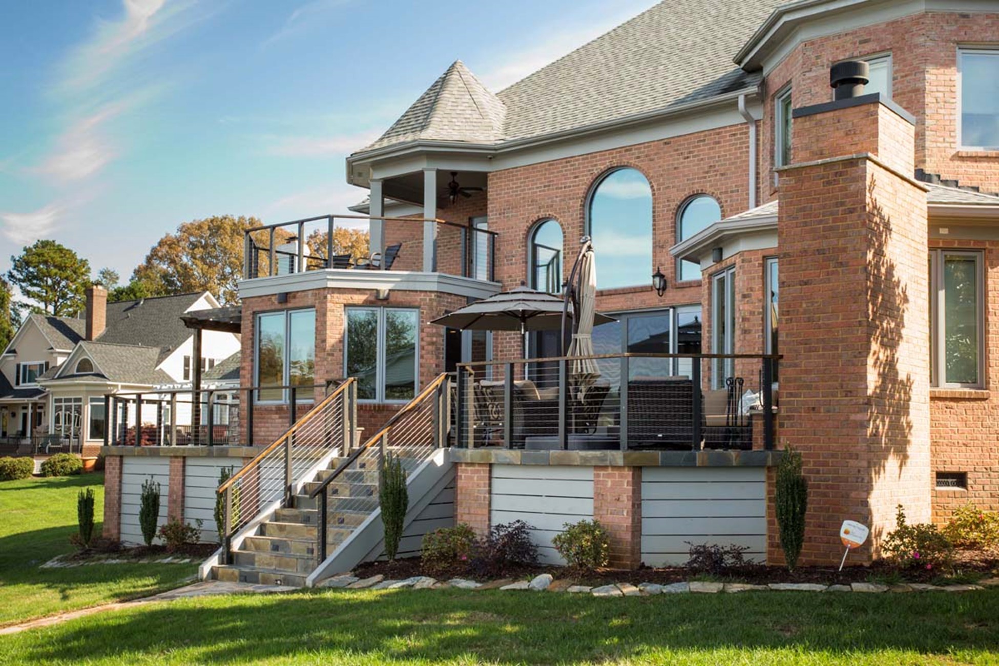 A two-story brick house with large windows and a balcony, featuring a patio with outdoor furniture and an umbrella, set in a grassy yard on a clear day.