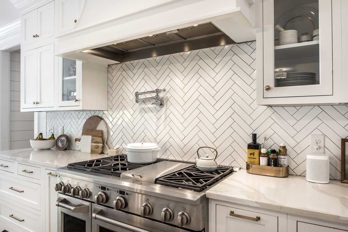 Modern kitchen with white cabinets and a herringbone tile backsplash. Features a stainless steel kitchen range, oven, and vent hood. Countertop holds a white pot, kettle, and kitchen essentials.