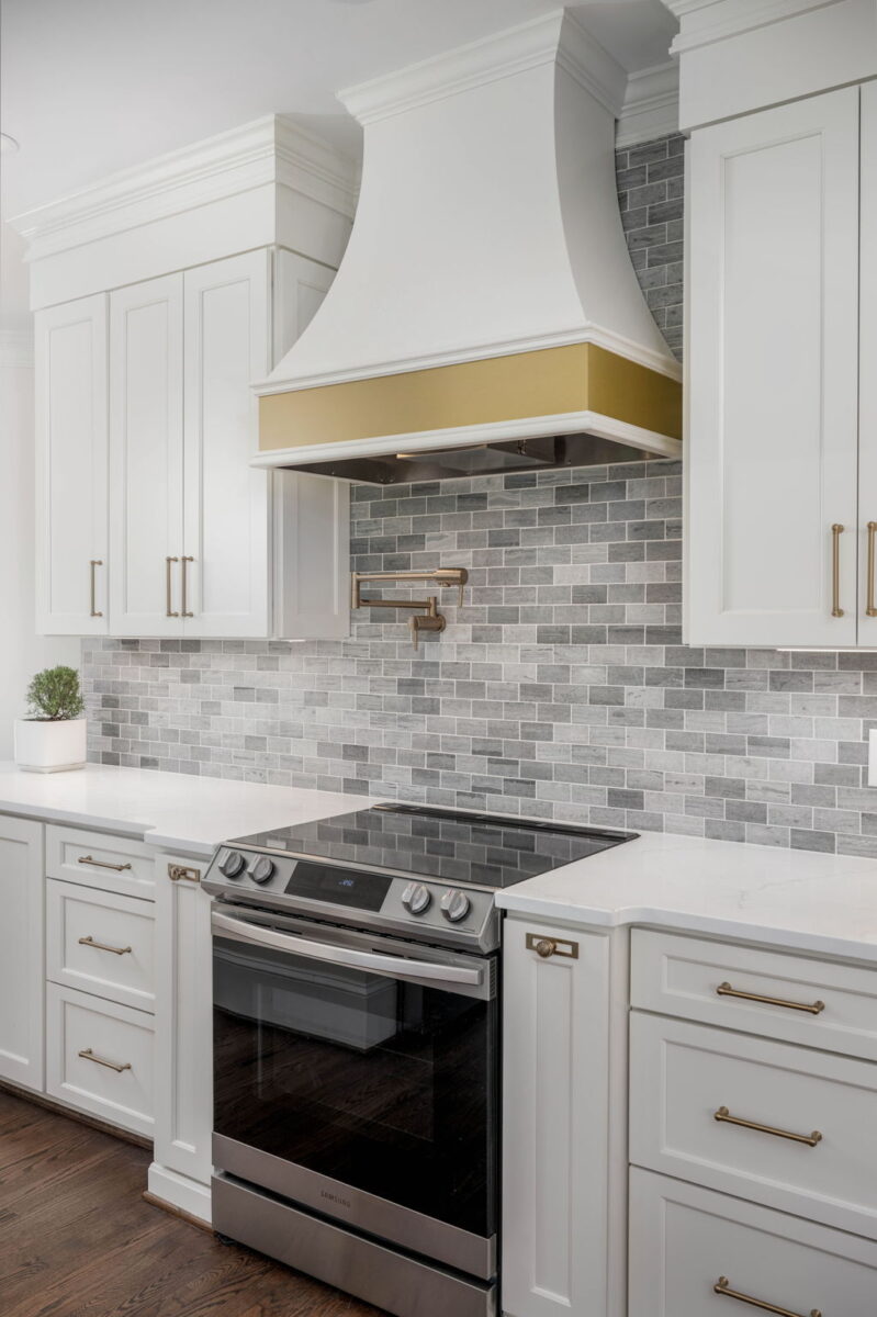 Modern kitchen with white cabinets, a gold-accented range hood, gray tile backsplash, and a black stovetop on a dark wood floor. Small plant on the left countertop.