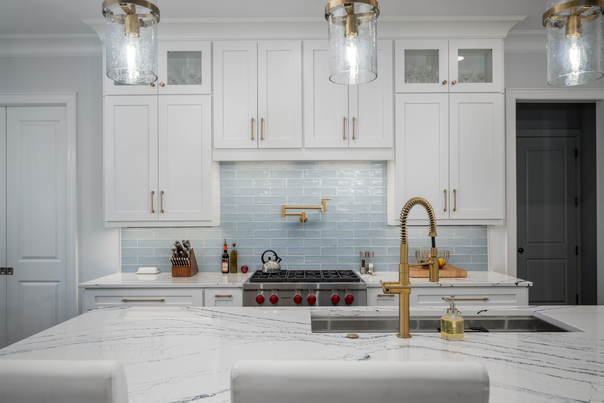 Modern kitchen with white cabinetry, a light blue tile backsplash, and stainless steel appliances. Features a marble island with a gold faucet and overhead glass pendant lights.