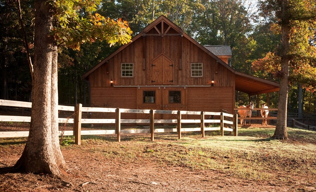 A wooden barn in Waxhaw NC with a pitched roof stands behind a fenced yard surrounded by trees in a sunlit forest setting.