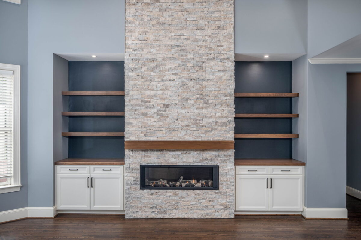 Modern living room with a central stone fireplace, flanked by dark shelves and white cabinets, against a blue-gray wall.