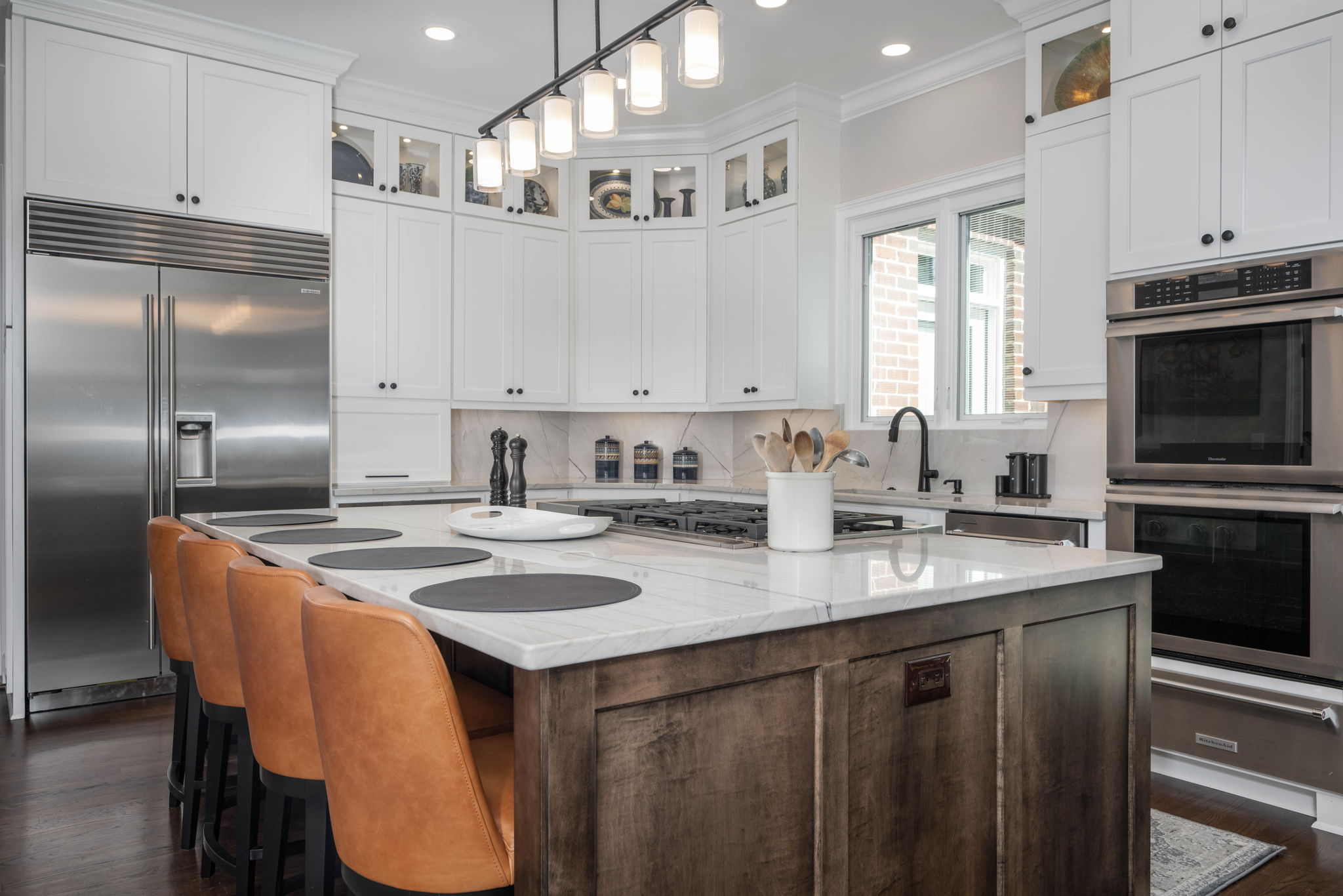 Modern kitchen with white cabinets, stainless steel appliances, a central island with brown leather stools, and a hanging light fixture.