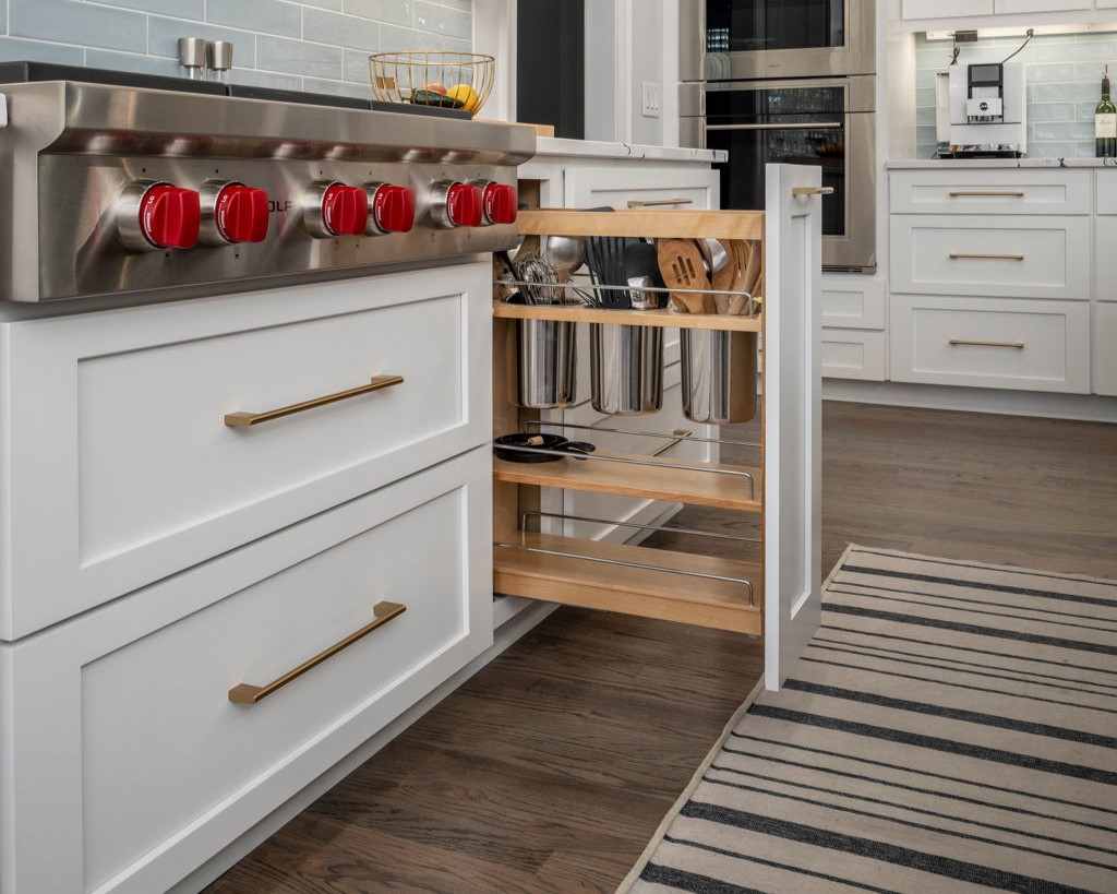 White kitchen cabinets features a slide-out drawer holding utensils. The countertop has red stove knobs, and a striped rug covers part of the wooden floor.