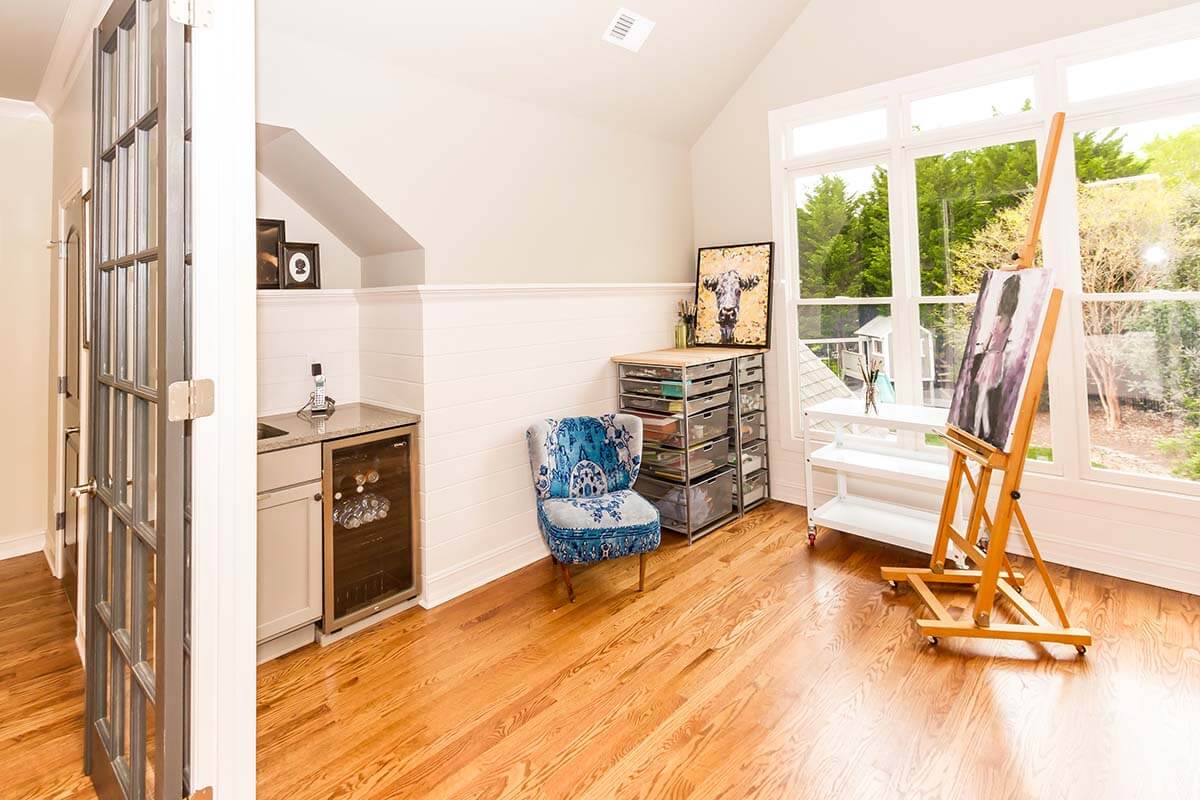 This type of home addition is a bright art studio with wooden floors, a blue patterned chair, easel, artwork, storage drawers, and a small kitchenette. Large windows offer a view of trees outside.