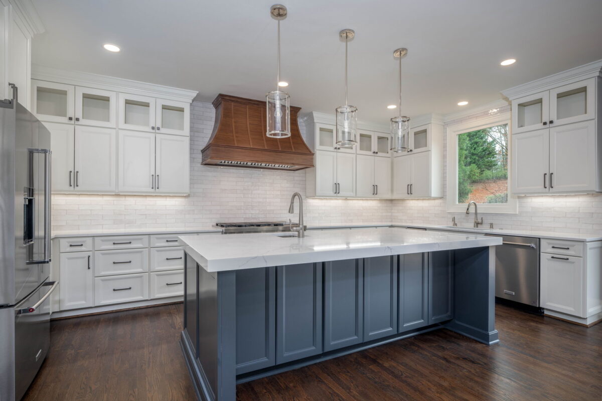 Modern kitchen remodel with white cabinets, dark wood floors, a large island with a gray base, pendant lights, and a wood vent hood above the stovetop.