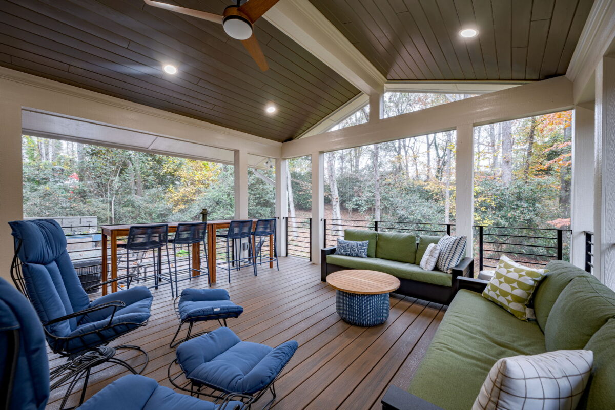 Covered outdoor patio with seating area, including sofas, chairs, and a round table. A ceiling fan is overhead, and a bar area with stools overlooks a wooded backyard—a perfect addition when considering adding a room to your house for relaxing or entertaining.