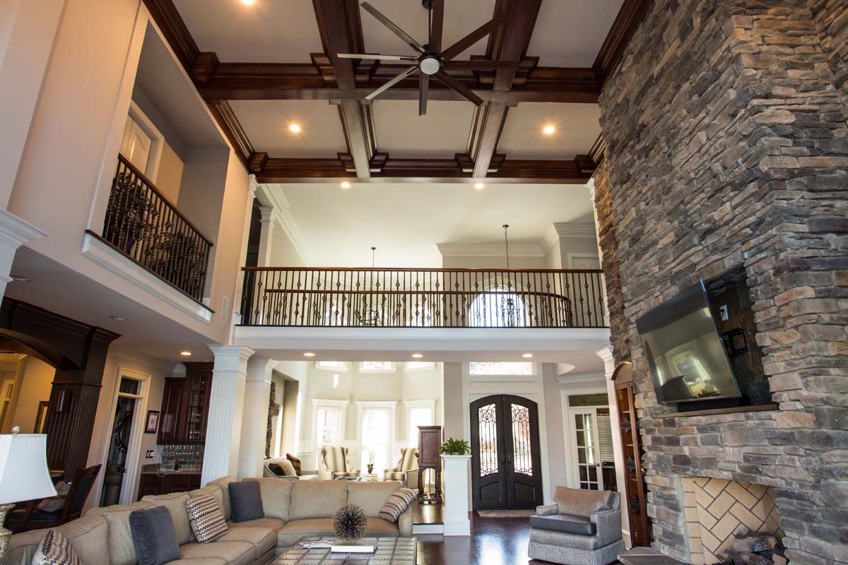 Considering adding a room to your house? This spacious living room features high ceilings, a large stone fireplace, wooden beams, a ceiling fan, a sectional sofa, and a charming balcony—perfect inspiration for your next home project.