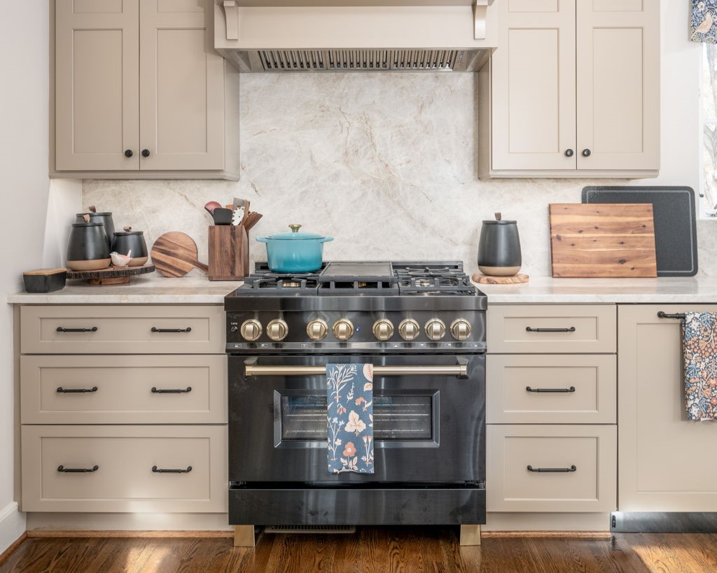 Modern kitchen with a black stove, beige cabinets, and a marble backsplash. A teal pot is on the stove, while various kitchen items are arrayed on the counters, showcasing professional home remodeling at its finest.