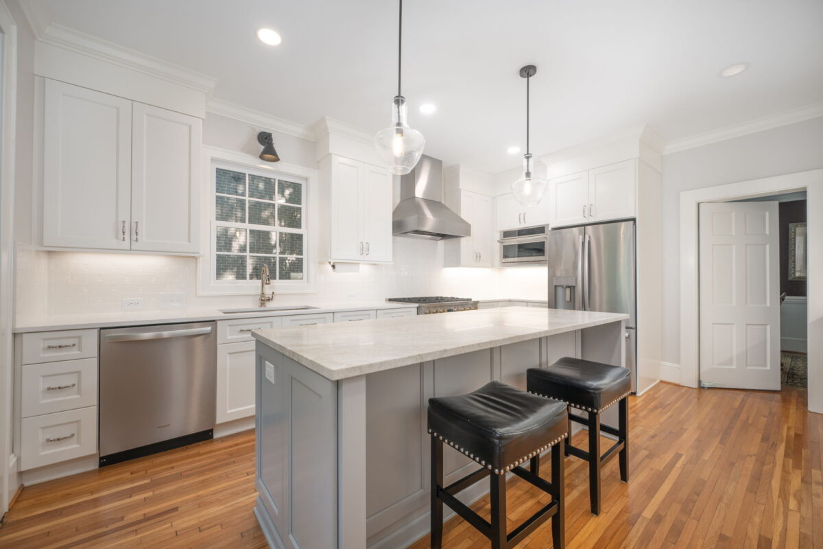 A modern kitchen with white cabinets, stainless steel appliances, and a large island with two black barstools on a hardwood floor. Pendant lights hang above the island.