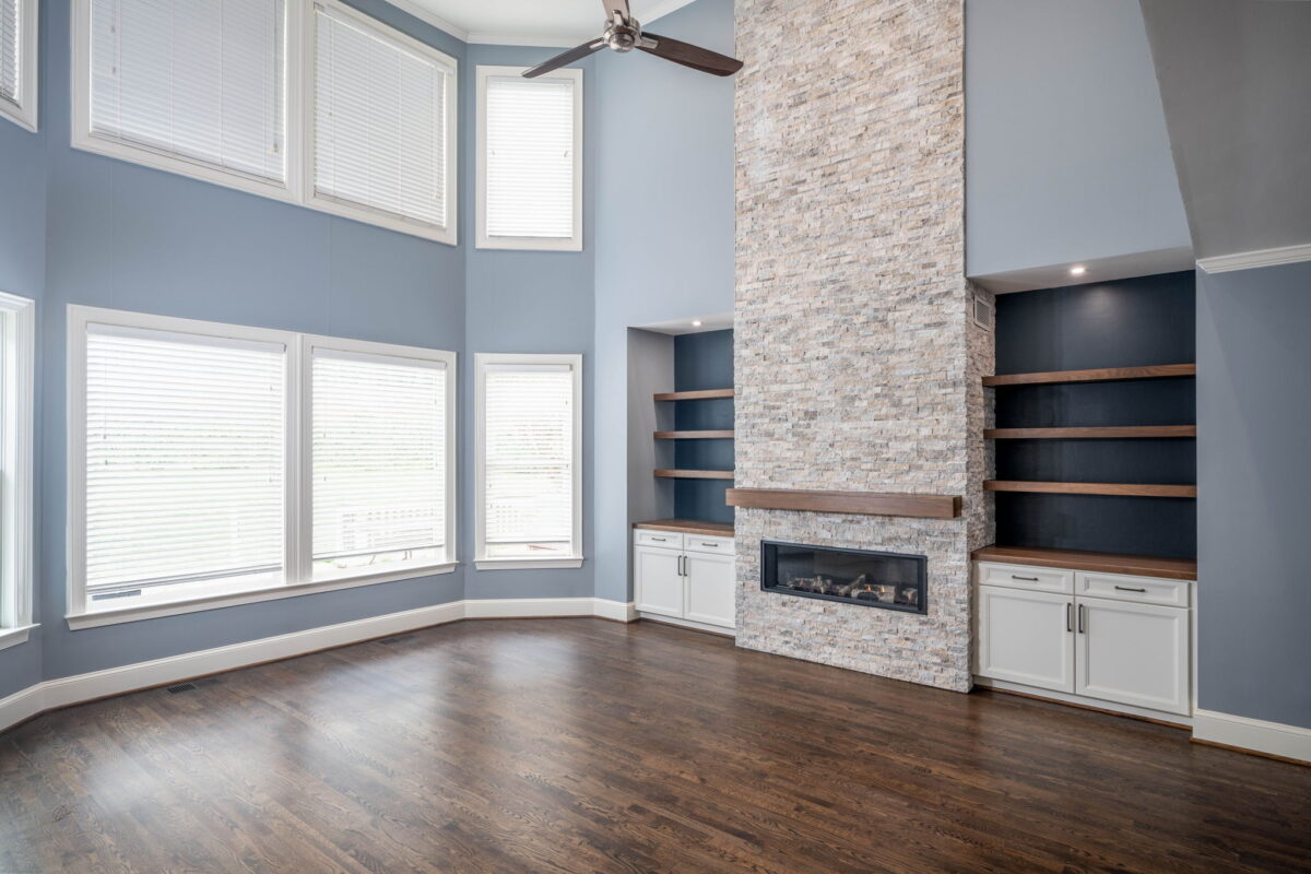 Spacious living room with high ceilings, stone fireplace, built-in shelves, large windows with blinds, and dark wood floors.