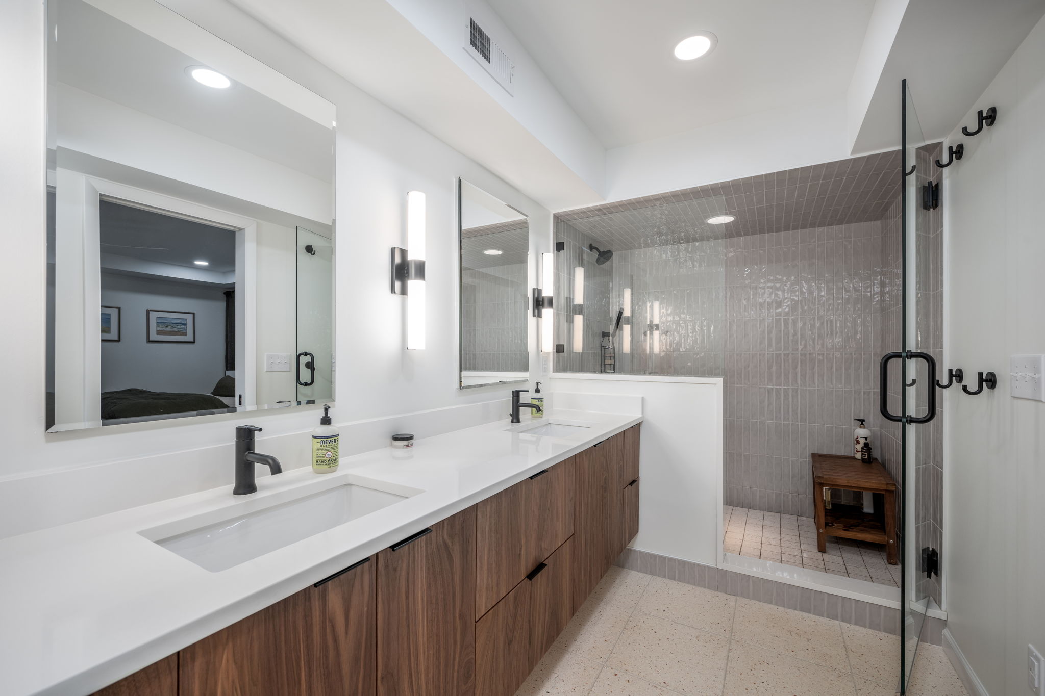 Modern bathroom with a double-sink vanity featuring wooden cabinets, wall-mounted lights, and a glass-enclosed shower with a tiled interior.