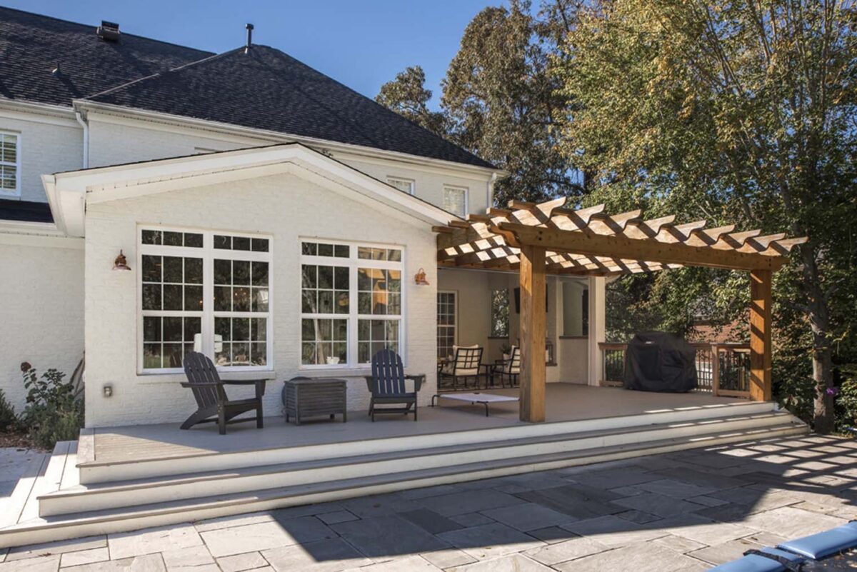 A white house with a covered patio and wooden pergola. There are chairs and a table on the patio, surrounded by trees.