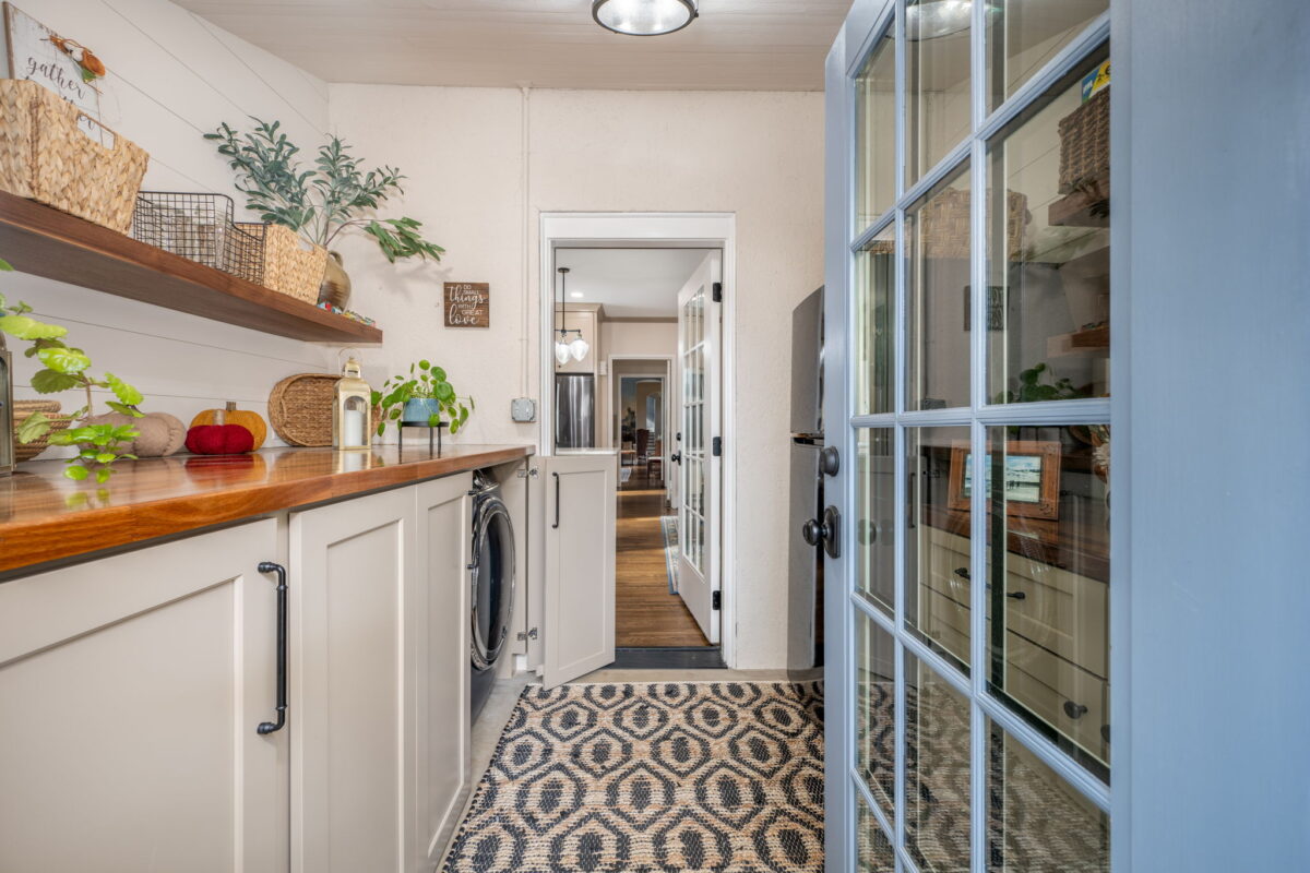 A bright laundry room with beige cabinets, wooden countertops, and a patterned rug. A glass door leads to a hallway. The room is decorated with plants and baskets.