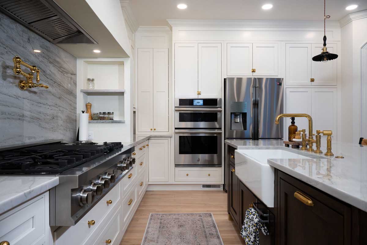 Modern kitchen with white cabinets, stainless steel appliances, a double oven, marble backsplash, and brass fixtures. Wooden flooring and a central island with a white countertop are visible.