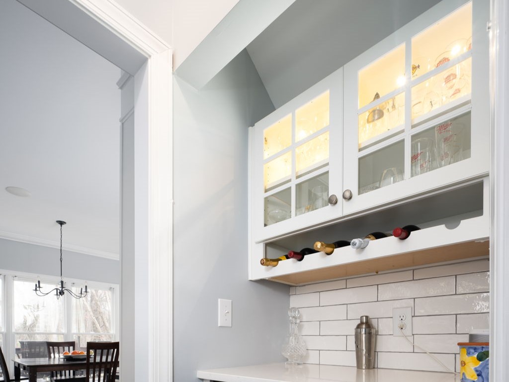 Modern kitchen corner with white cabinets, wine rack, glass shelves, and countertop. Subtle lighting accents contents. Dining area with wooden table and chairs visible in the background.