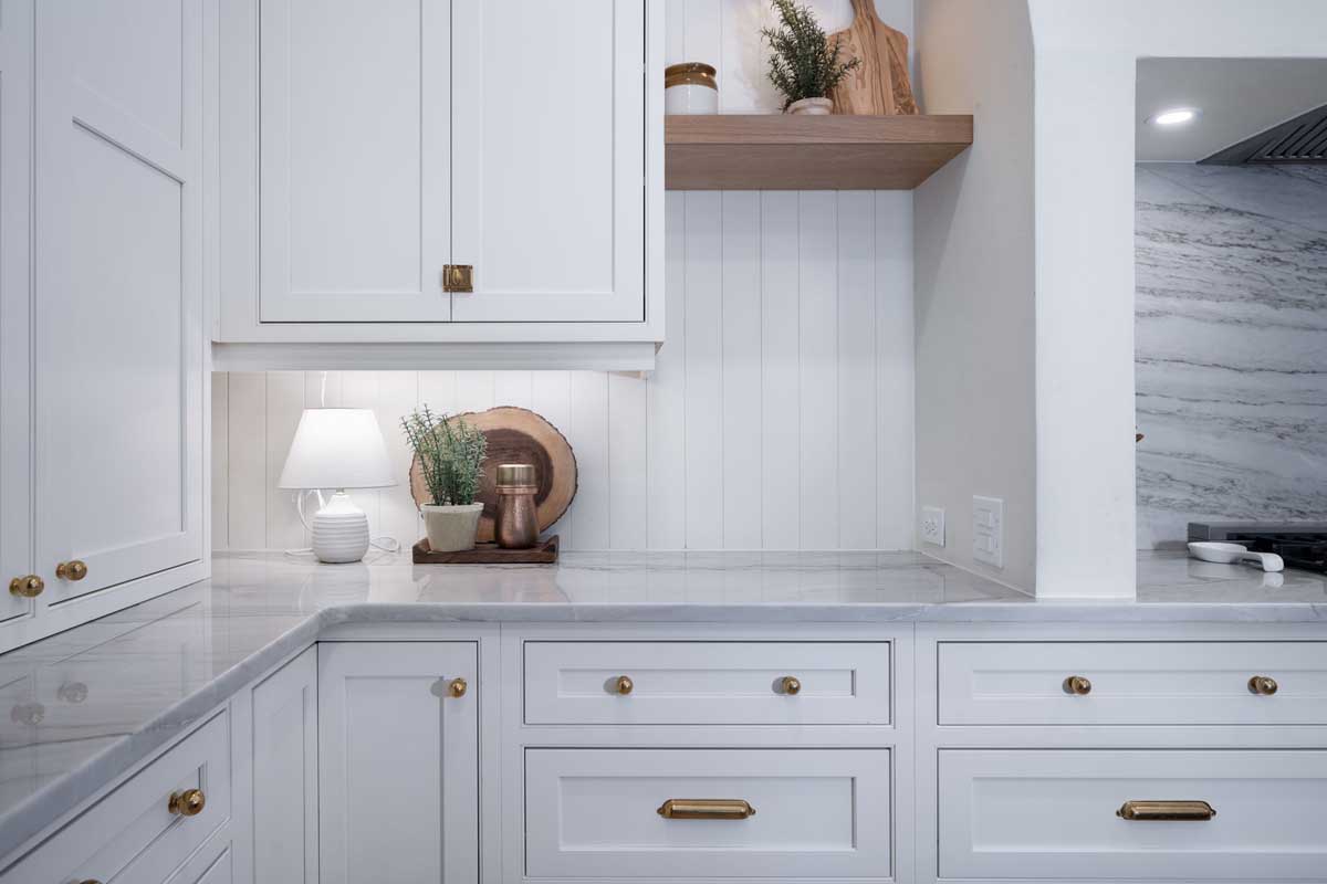 A kitchen with white cabinets, brass handles, a marble countertop, and wooden accents. A small lamp and plants are on the counter under a wooden shelf.