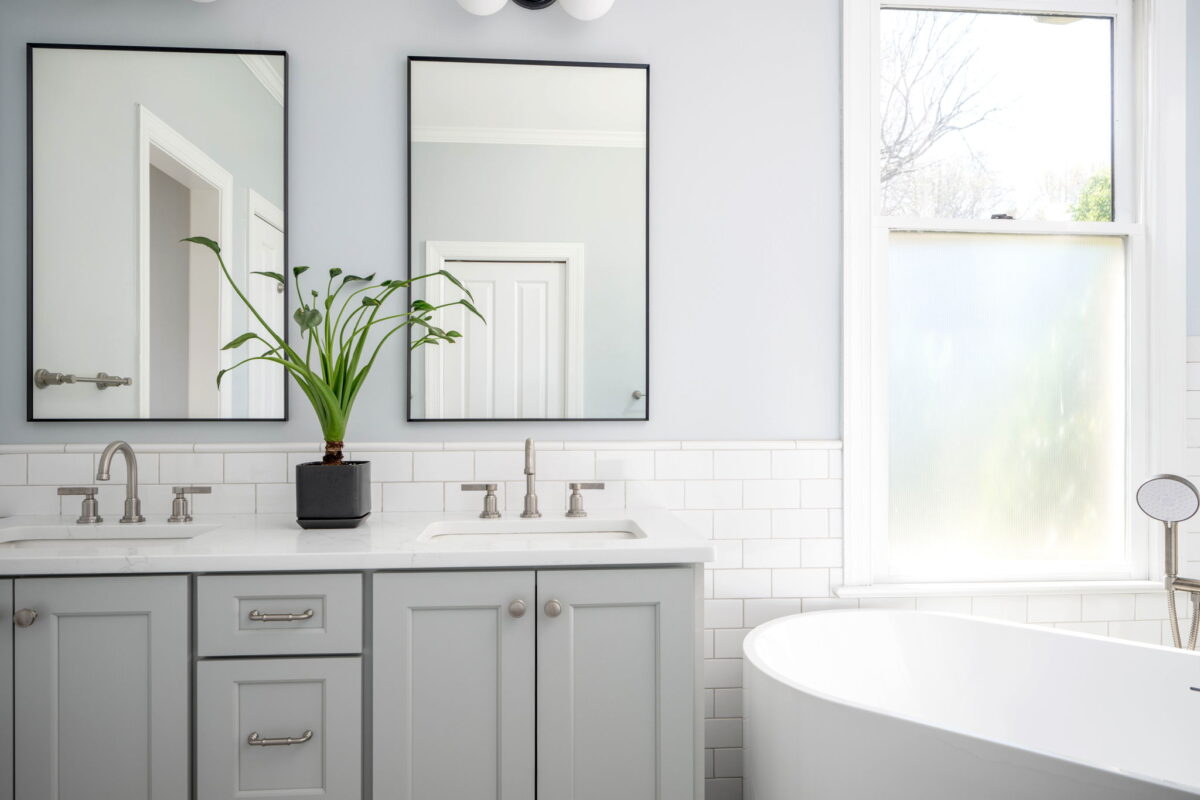 Modern bathroom with a double-sink vanity, two mirrors, a potted plant, and a white freestanding bathtub next to a frosted window.