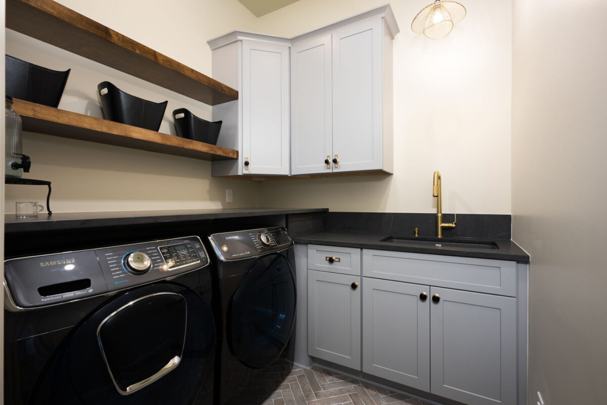 Modern laundry room with black washer and dryer, white cabinets, open shelves with bins, black countertop, gold faucet, and herringbone tile floor.