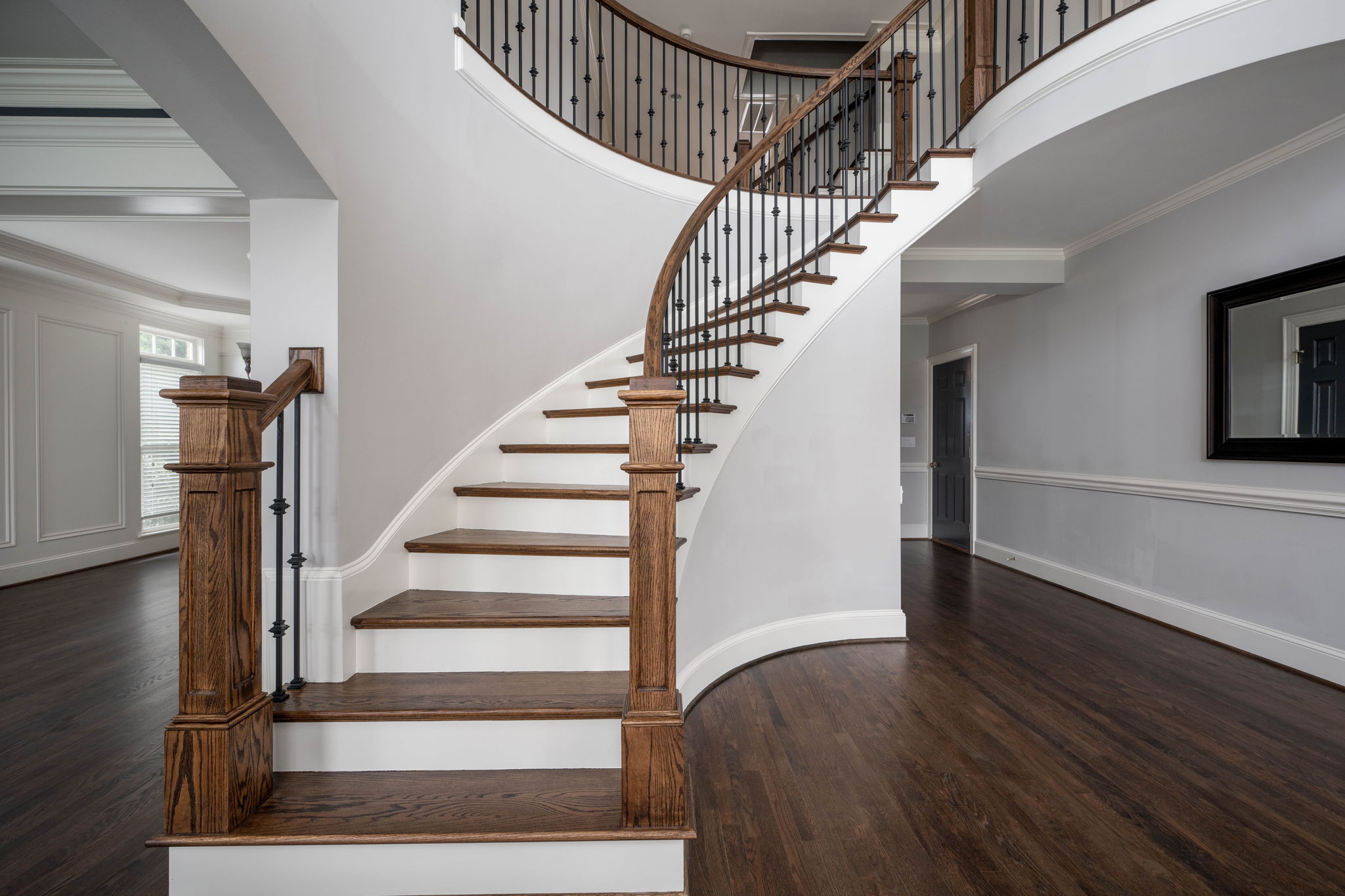 Curved wooden staircase with dark railing in a modern, open foyer with white walls and dark hardwood floors.